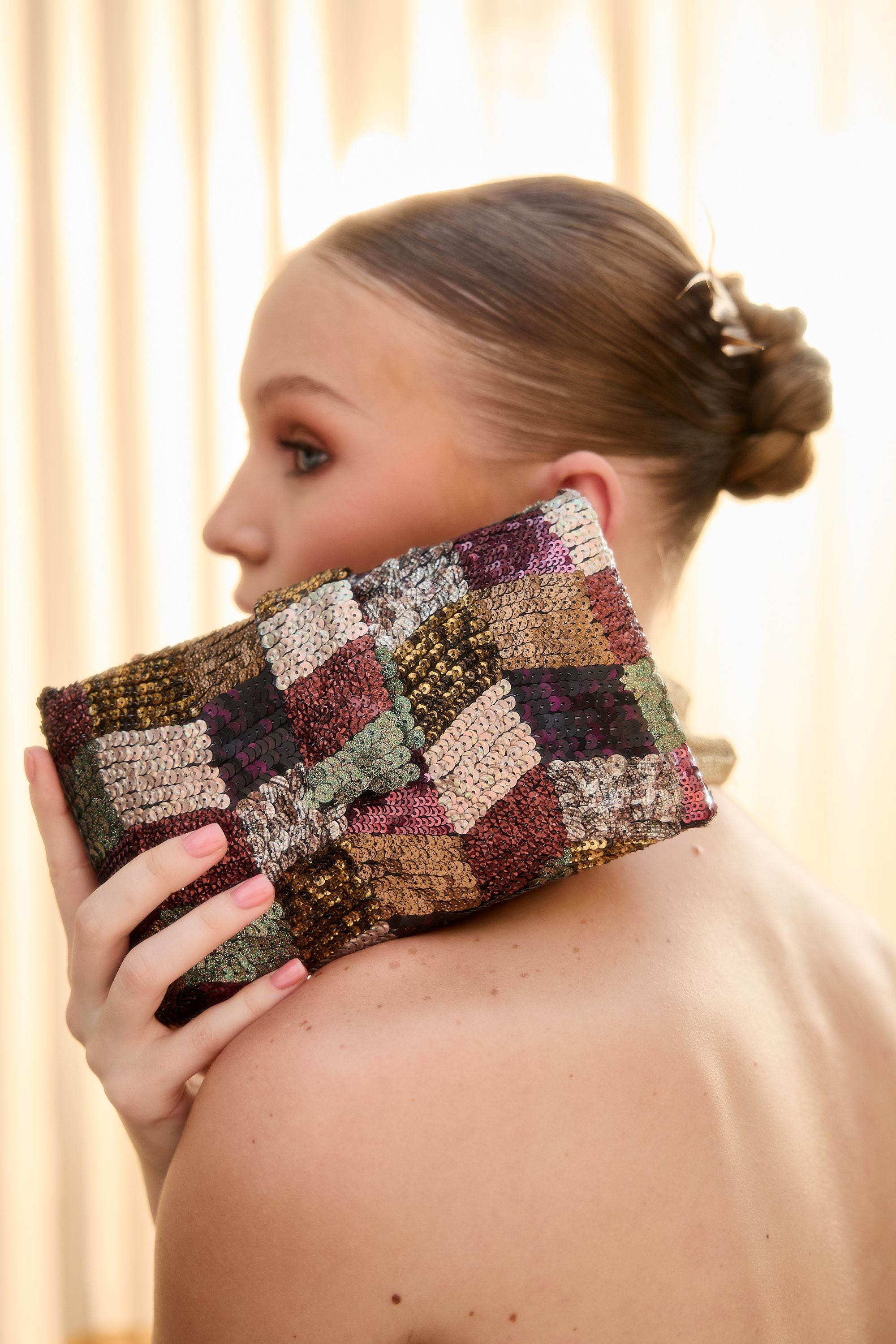 A woman holds a square patchwork clutch bag against her shoulder. The clutch features a multicolor geometric pattern with metallic gold, green, purple, and brown textured squares.
