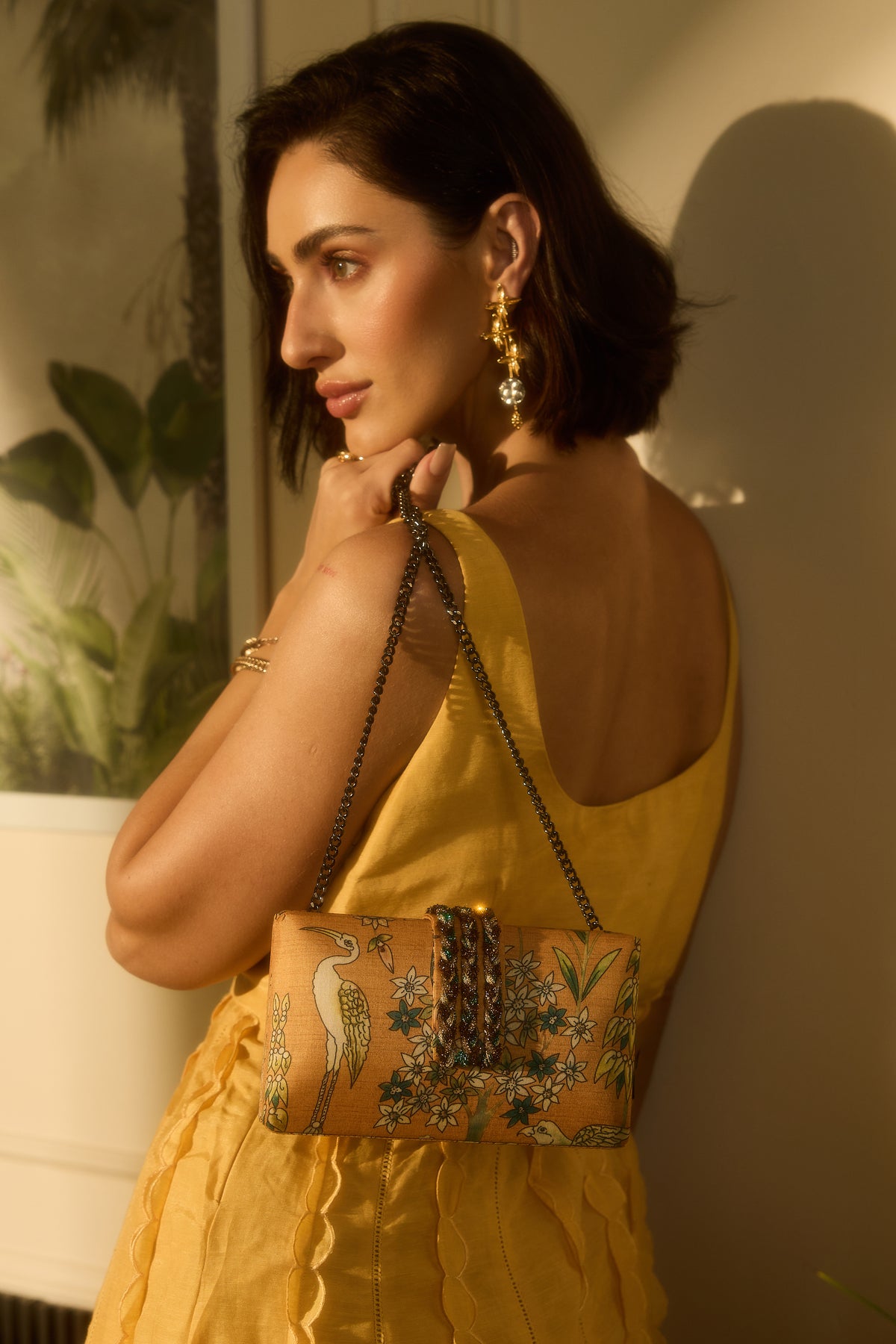 A woman in a yellow dress carries a tan leather clutch bag with a detailed bird and floral print, set against a warm, softly lit indoor background.