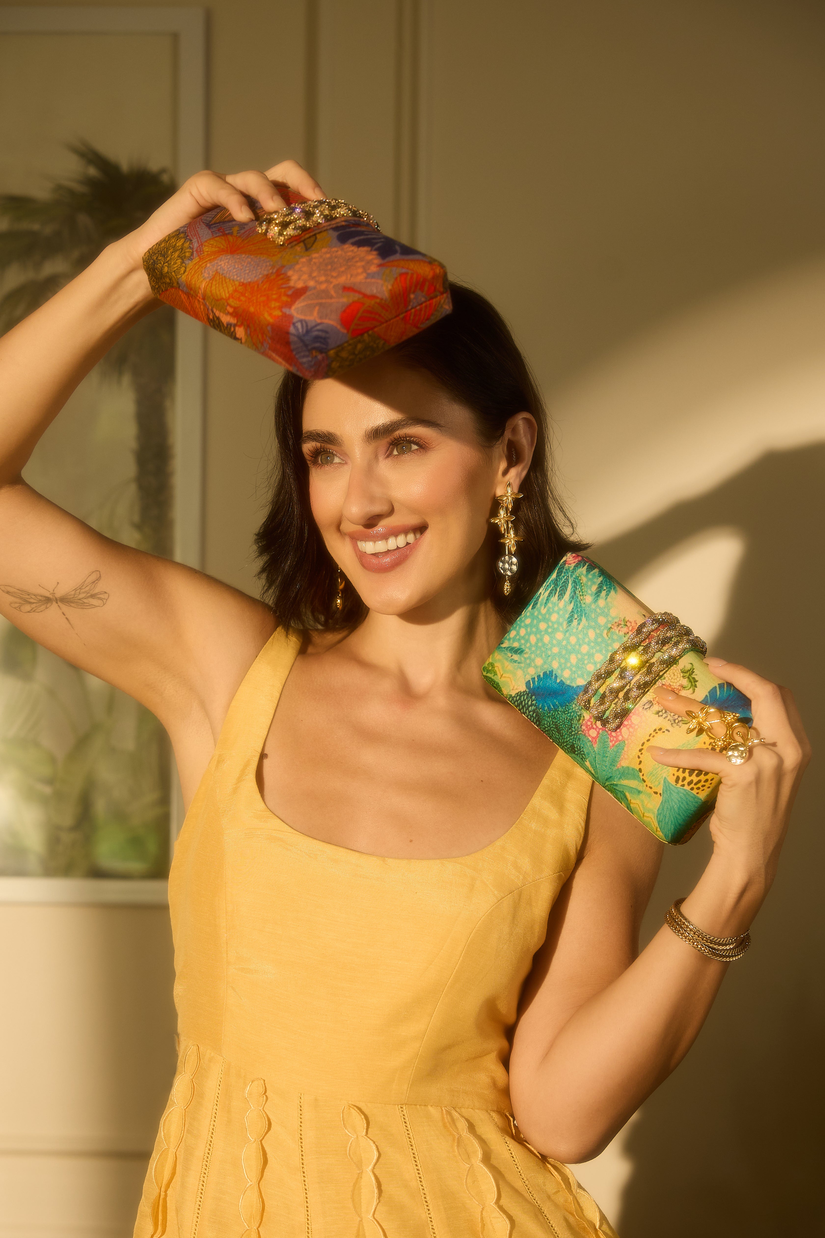 A woman in a yellow dress holds two colorful patterned clutch bags, one orange-red and one green-blue, against a warm-toned indoor background with dramatic shadows.