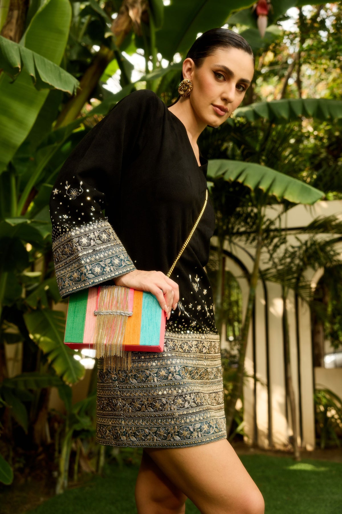A woman wearing a black embroidered mini dress with ornate metallic detailing on the sleeves and hem, holding a colorful fringe clutch bag, posed outdoors in front of lush green tropical foliage.