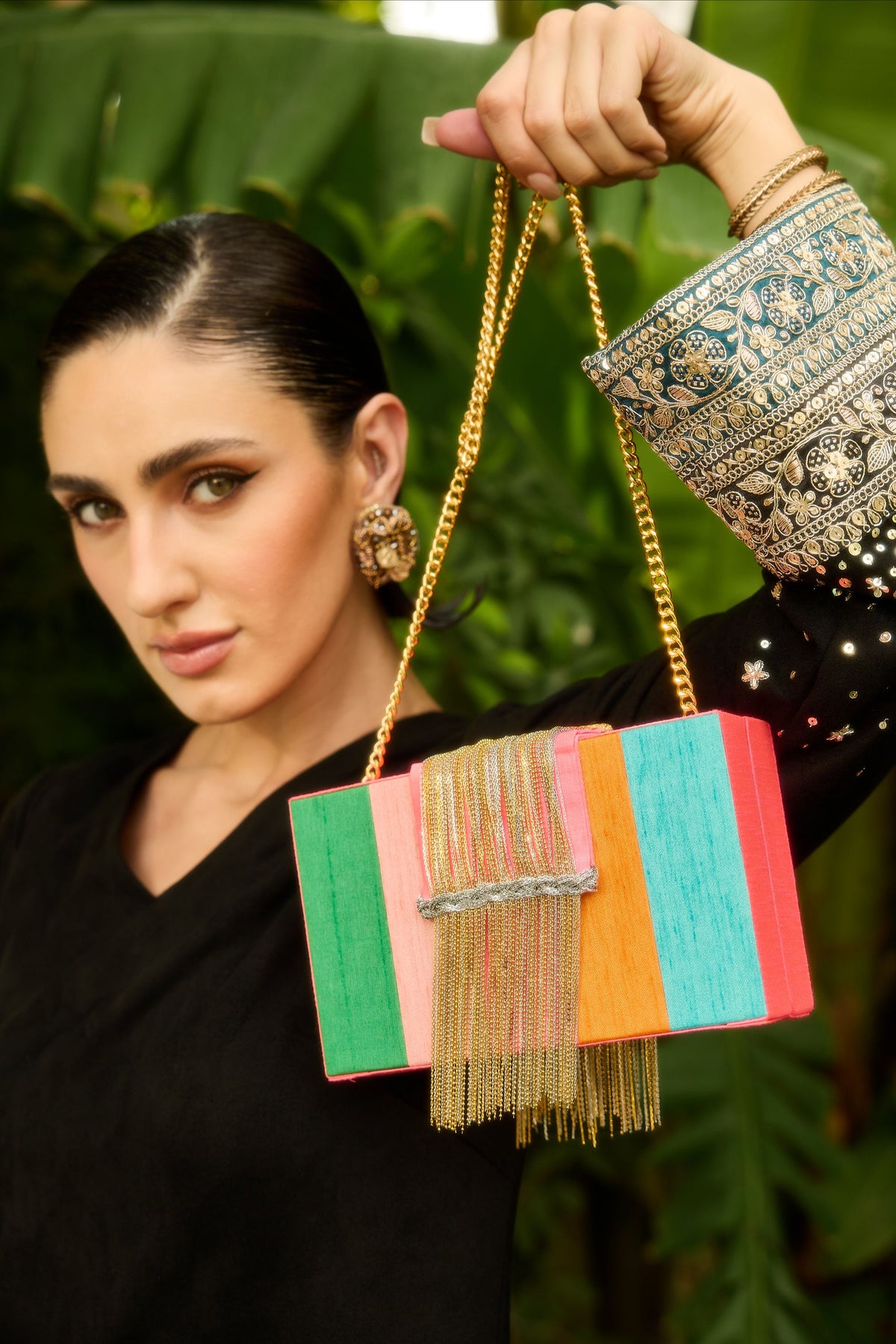 A woman in a black outfit holds up a colorful rectangular clutch bag with gold chain straps and gold fringe. The clutch features vertical panels in green, pink, orange, and turquoise, with a silver beaded band and gold fringe.