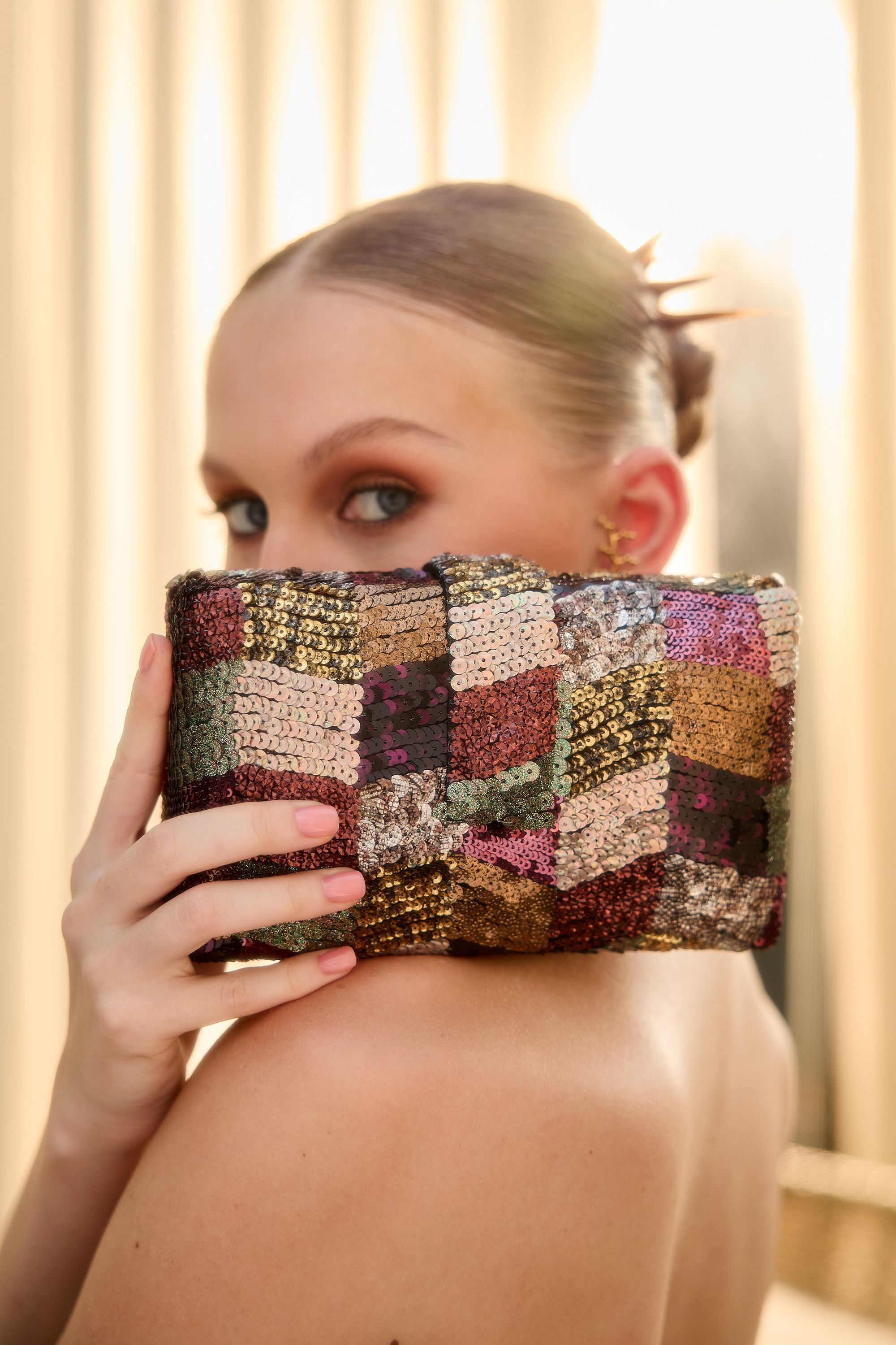 A woman holds a Antique Zigzag Kitsch Clutch in front of her face. The clutch features a multicolor patchwork design with shimmering metallic and textured squares in gold, bronze, pink, purple, and green.