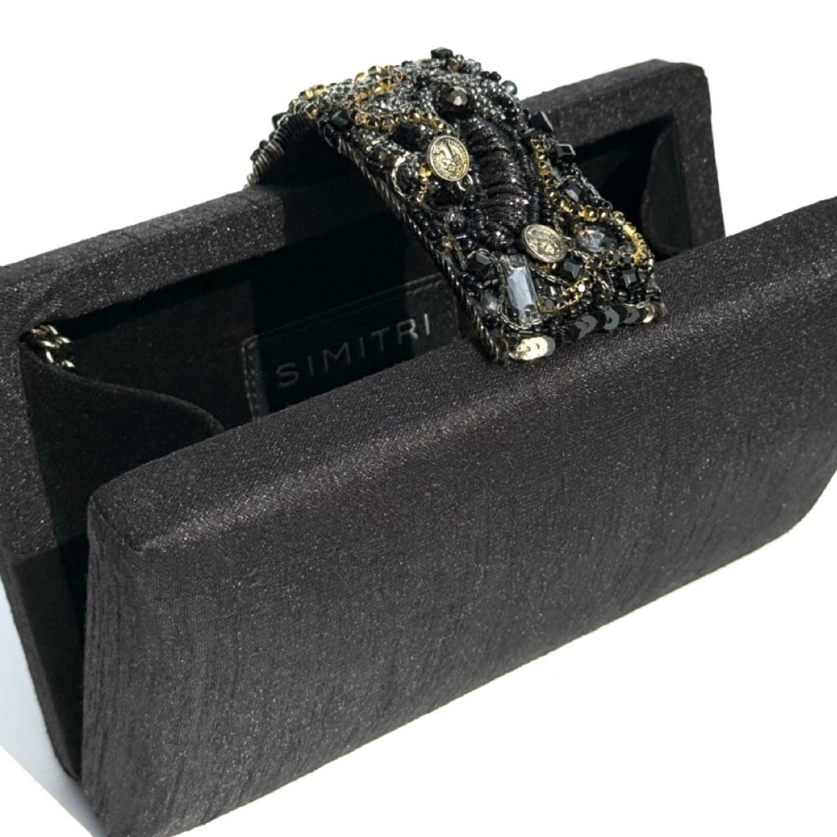 A black textured clutch bag with a wide, heavily embellished handle featuring black beads, gold-toned coins, and clear rhinestones. The brand name 'SIMITRI' is visible on the interior label.