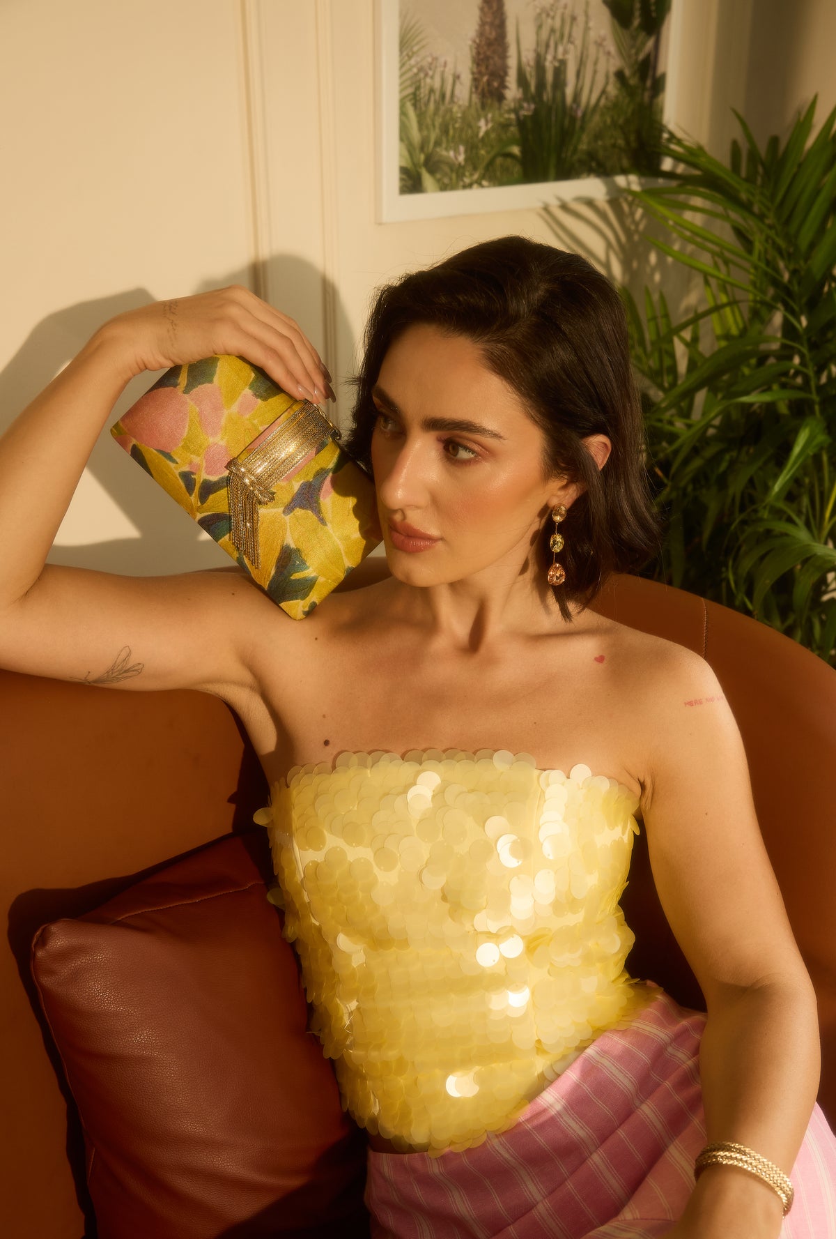 A woman posing on a brown leather couch wearing a yellow sequined strapless top and a pink plaid skirt, holding a yellow patterned clutch.
