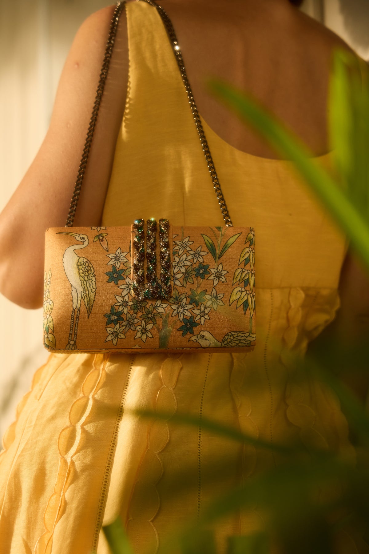 A woman in a yellow dress carries a small rectangular clutch bag with a floral and bird print. The clutch has a gold-tone chain strap and a decorative clasp, photographed in warm indoor lighting with blurred green leaves in the foreground.