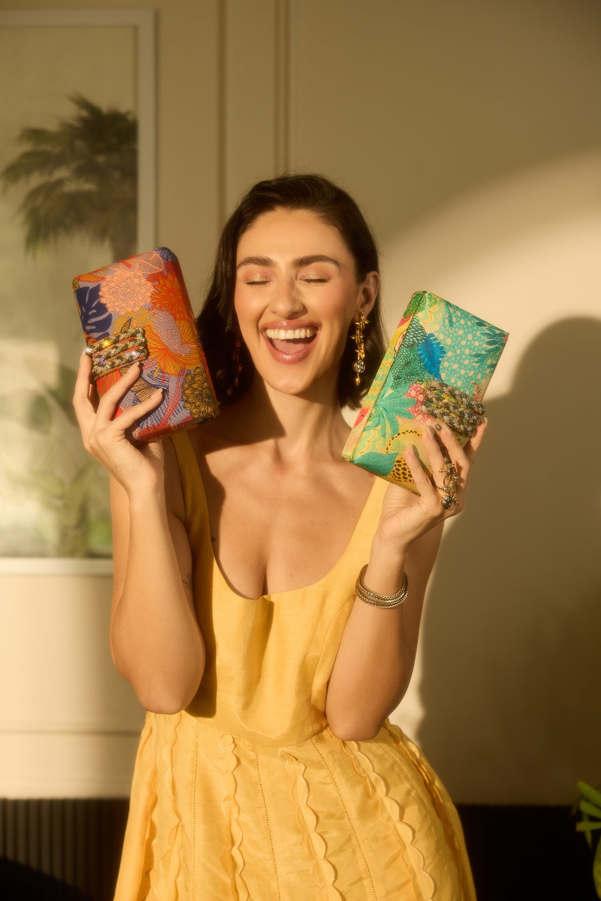 A woman in a yellow dress holds up two colorful patterned clutch bags, one orange and blue, and one green and blue, in a warmly lit indoor setting.