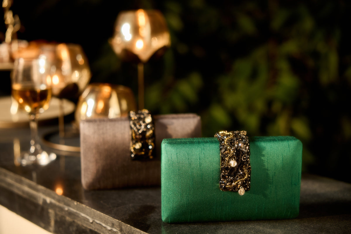 A green satin clutch purse with an ornate gold and black beaded strap closure, displayed on a dark table next to two matching satin evening bags in a warm, dimly lit setting.